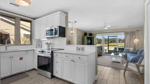 Una cocina moderna con armarios blancos, horno empotrado y microondas. Hay una zona de comedor con sofá y una mesa baja.