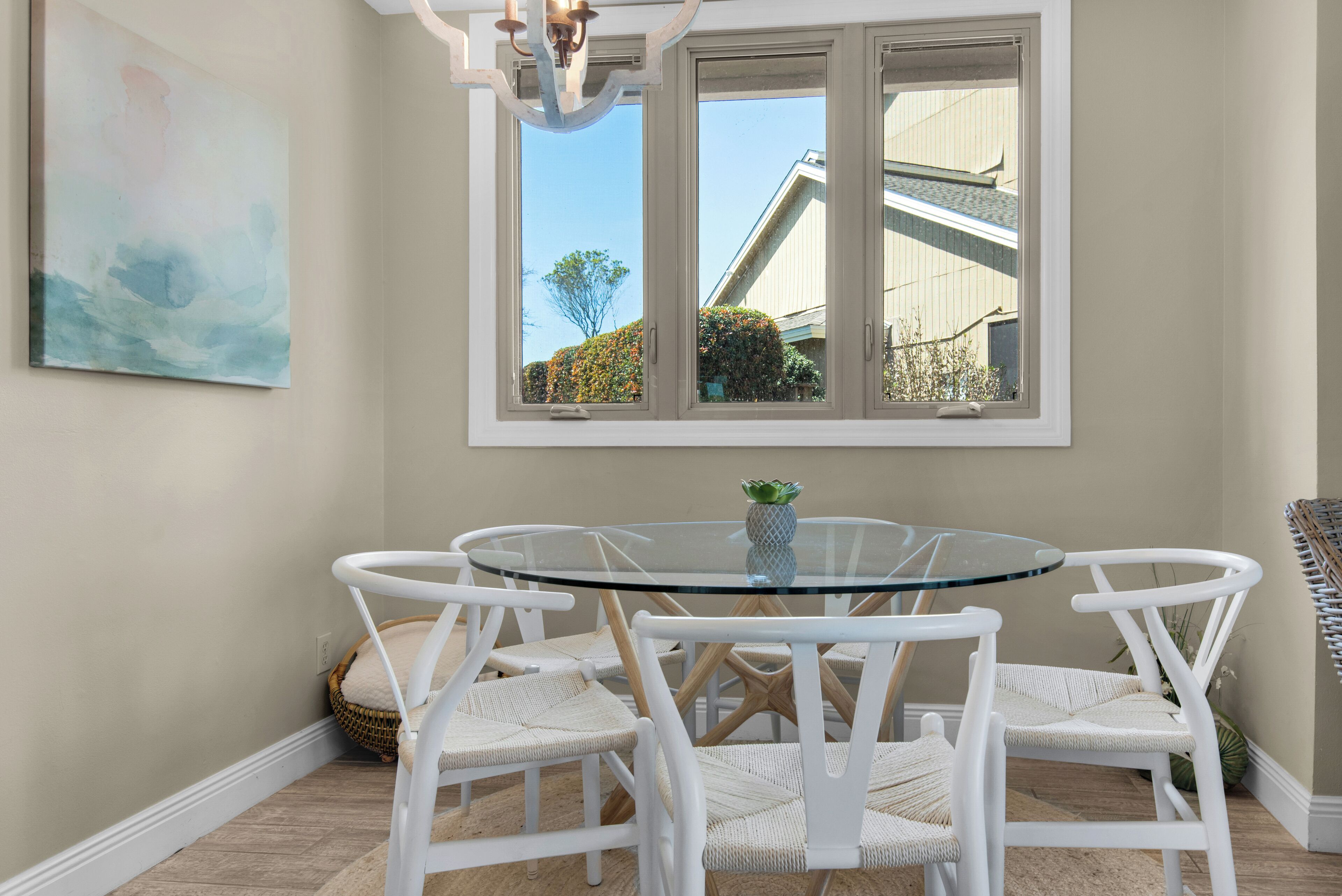 A dining area with a round glass table surrounded by white wicker chairs, a painting on the wall, and a window with a view of a building and trees.