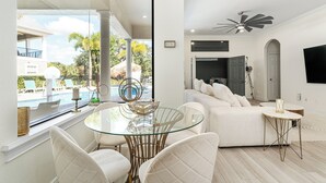 A modern living room with a glass dining table, white sofas, and a view of a pool and outdoor furniture. - Rare Extended Pool Deck & Oversized Spa | Arcade Room | Kids Bedrooms (Kissimmee)