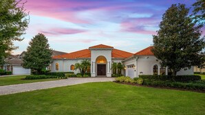 A large, single-story house with a red-tiled roof, a central entrance with an arched doorway, and a well-maintained lawn. - Rare Extended Pool Deck & Oversized Spa | Arcade Room | Kids Bedrooms (Kissimmee)