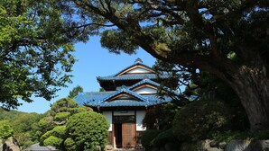 A traditional Japanese building with a tiled roof, surrounded by lush greenery and large trees. - Kougetsu Sanso (Hachioji)