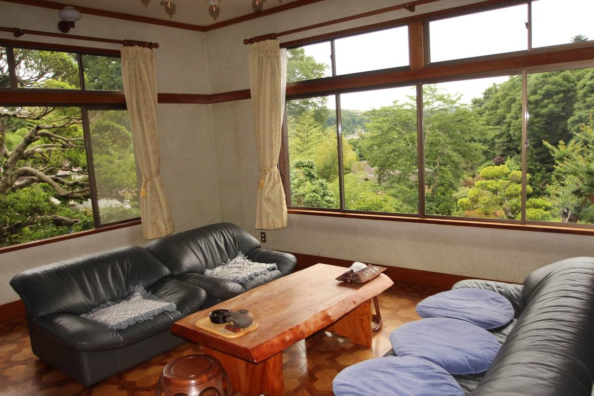 A room with a wooden coffee table, a black leather sofa, and a view of a lush green landscape through large windows.