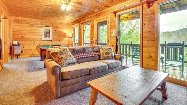 A cozy cabin interior with a wooden ceiling, a sofa with patterned cushions, a wooden table, and a view of the outdoors through large windows. - 2-Tier Deck & Hot Tub: Quiet Sevierville Cabin (Sevierville)