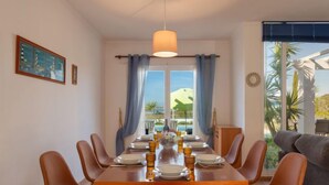A dining area with a wooden table set for six, surrounded by brown chairs, a hanging light fixture, and a view of a pool and mountains through large windows. - Villa Blue Ocean by Interhome (Gale)