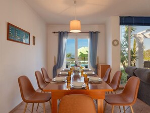 A dining area with a wooden table set for six, surrounded by brown chairs, a hanging light fixture, and a view of a pool and mountains through large windows.