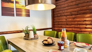 Ein Holztisch mit einer Schüssel Brot, einer Saftflasche und Gewürzen. Grüne Stühle stehen darum, und an der Seite befinden sich Blumentöpfe.