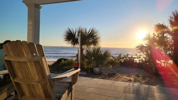 Una silla de madera y una mesita bajo una pérgola con vista a la playa al atardecer.