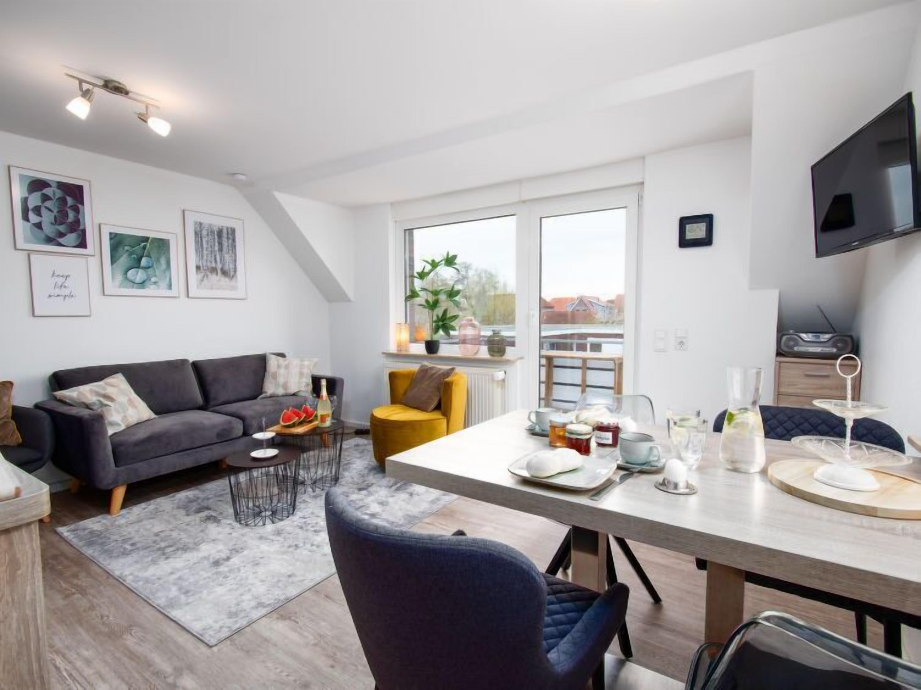 A modern living room with a grey sofa, a yellow armchair, and a dining table set with glasses and a bottle.