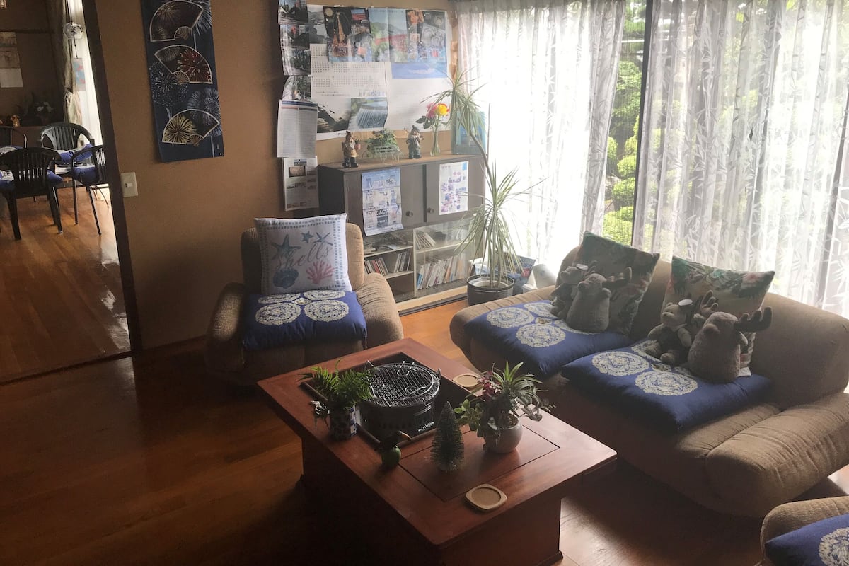 A living room with a wooden coffee table, two sofas with patterned cushions, a bookshelf, and a large window with sheer curtains.