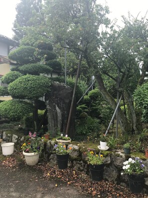 A garden with various potted plants and trees, including a large rock and a small white house-like structure. - Kizuna no Ie (Uozu)