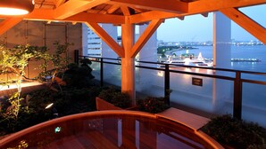 A wooden deck with a pool, surrounded by plants and overlooking a body of water with boats and buildings in the background. - Yokohama Minamimirai Manyo-Club (Yokohama)