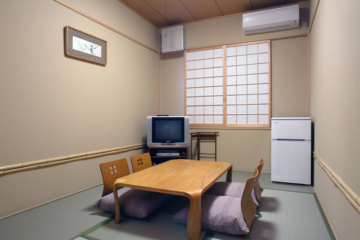 A small room with tatami flooring, a wooden table, a television, a refrigerator, and a sliding door.
