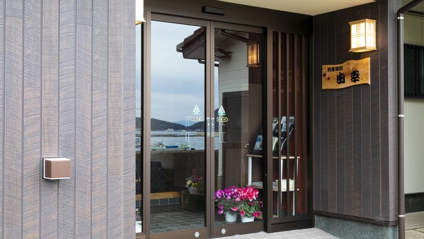 A modern building entrance with a glass door, a wooden sign with Chinese characters, and a small outdoor seating area with flowers. - Yoshikou (Takahama)