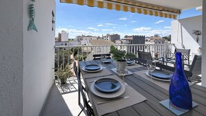Un balcon avec une table dressée pour deux personnes, une plante en pot et un vase bleu.