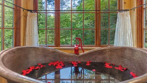 Eine rustikale Badewanne mit roten Blütenblättern im Wasser, einem roten Wasserhahn und Blick auf einen Wald durch große Fenster.