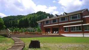 A red brick building with a flat roof and large windows, a wooden deck with chairs, and a grassy area in front. - Sora no Mori Hotel Hanagori (Kami)