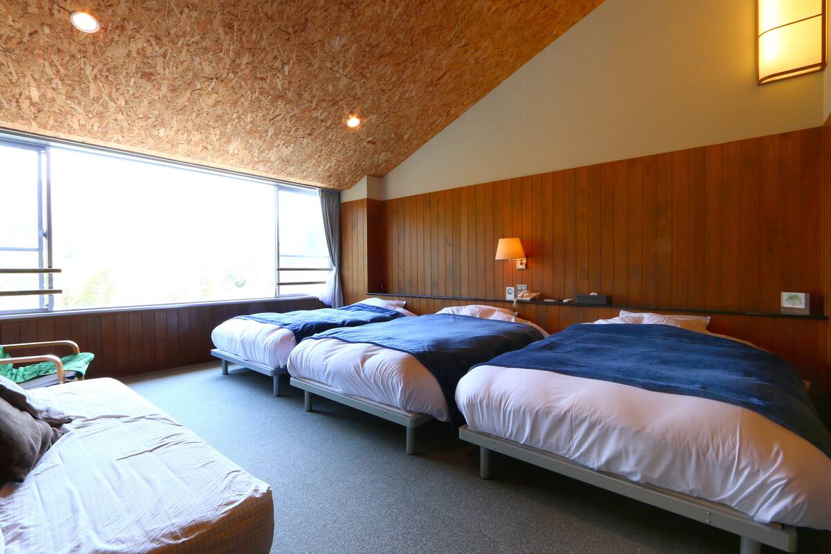 A room with three beds, wooden paneling on the walls, a window with curtains, and a bedside lamp.