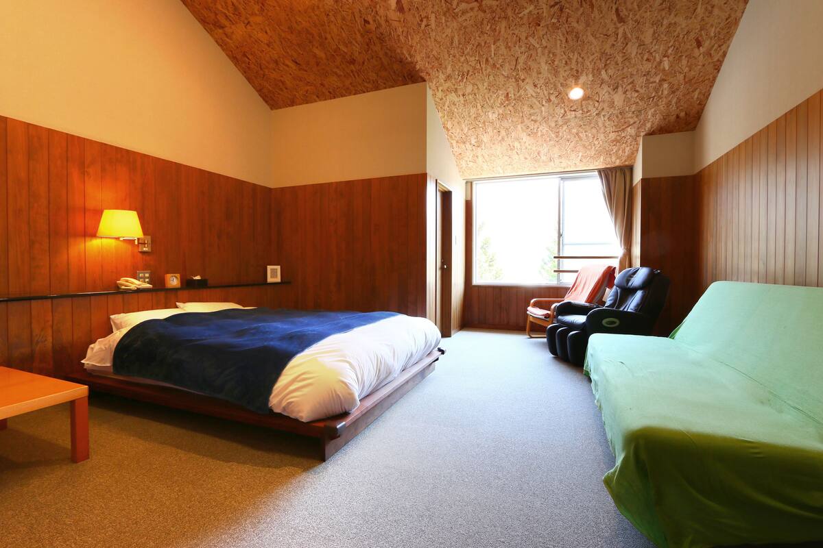A bedroom with a wooden ceiling, a bed with a blue blanket, a green sofa, a massage chair, and a bedside table.