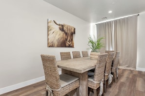 A dining area with a wooden table and wicker chairs, a large animal print artwork on the wall, and a potted plant.