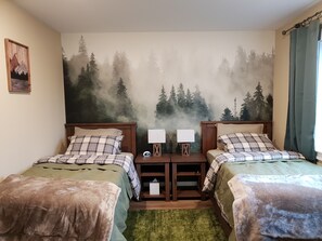 Un dormitorio con dos camas, una mesita de noche con lámparas, un mural de un bosque en la pared y una cortina verde.