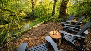 A forested area with a small stream, wooden chairs, and a lantern on a tree stump.