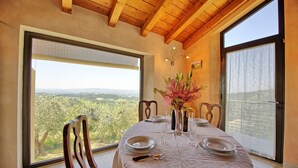 A dining area with a table set for four, large windows offering a view of the countryside, wooden ceiling beams, and a vase with flowers on the table. - Wonderful private villa with pool, A/C, WIFI, balcony and panoramic view, close to Greve In Chia... (Tavarnelle Val Di Pesa)