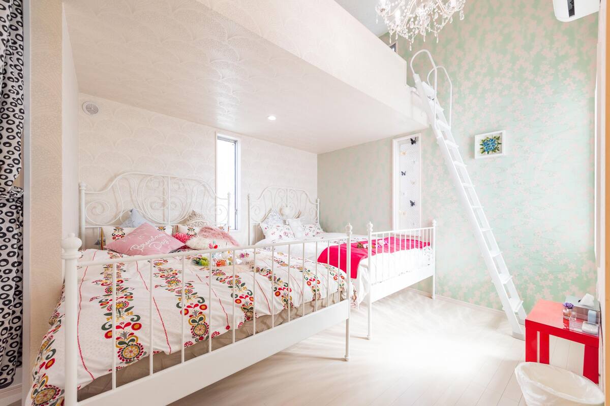 Une chambre avec un lit en métal blanc, une literie à motifs floraux, un plaid rouge et une échelle appuyée contre le mur.