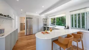A modern kitchen with a central island, wooden flooring, and large windows offering a view of greenery. - Noosa Escape 2BR Ocean Views, Walk to Beach (Noosa Heads)