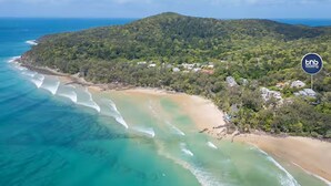 An aerial view of a coastal area with a beach, clear turquoise waters, and a forested hill. - Noosa Escape 2BR Ocean Views, Walk to Beach (Noosa Heads)