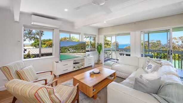 A spacious living room with a white sofa, striped armchairs, a wooden coffee table, and a TV. Large windows offer views of the outdoors. - Noosa Escape 2BR Ocean Views, Walk to Beach (Noosa Heads)