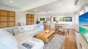 A spacious living room with a large sofa, a wooden coffee table, and a TV on the right. There is a dining area with chairs and a table in the background. - Noosa Escape 2BR Ocean Views, Walk to Beach (Noosa Heads)