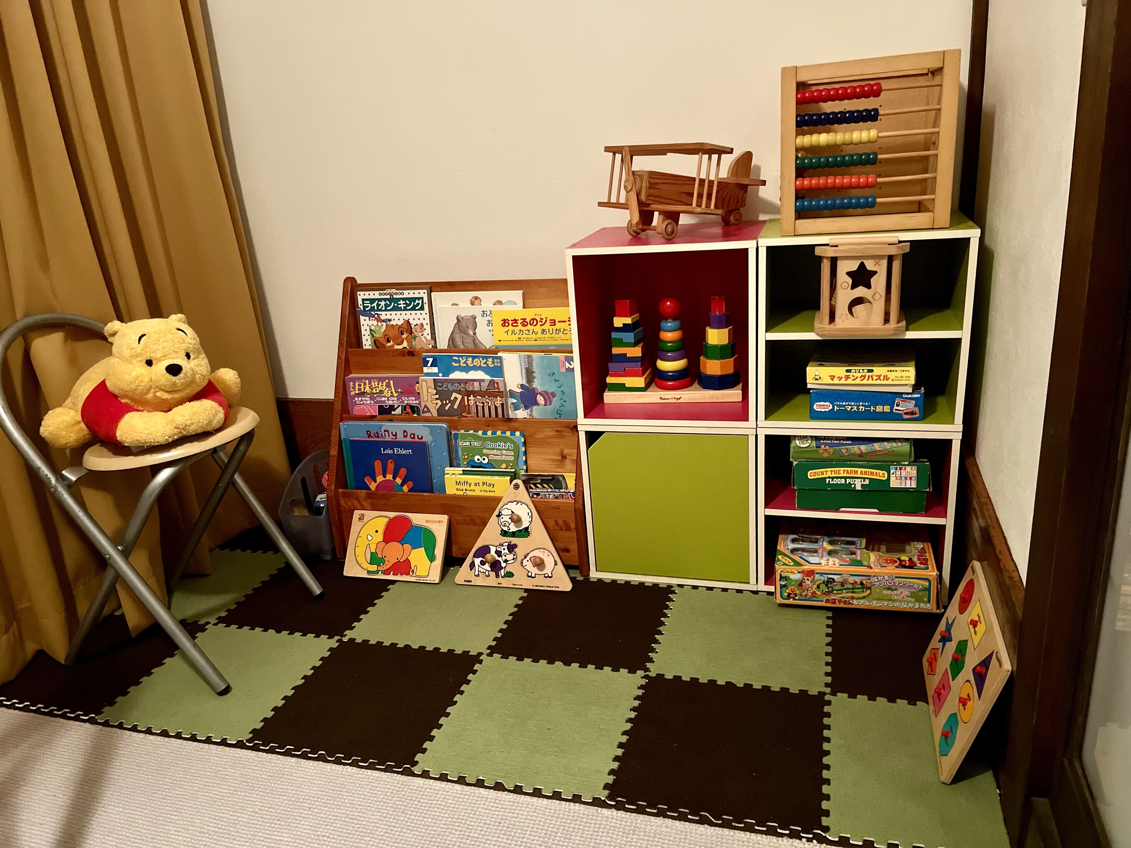 Zona de juegos infantiles con un osito de peluche en una silla, un estante de madera con libros y juguetes, y un suelo a cuadros.