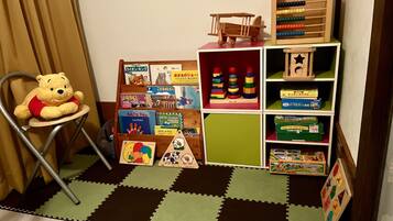 Zona de juegos infantiles con un osito de peluche en una silla, un estante de madera con libros y juguetes, y un suelo a cuadros.
