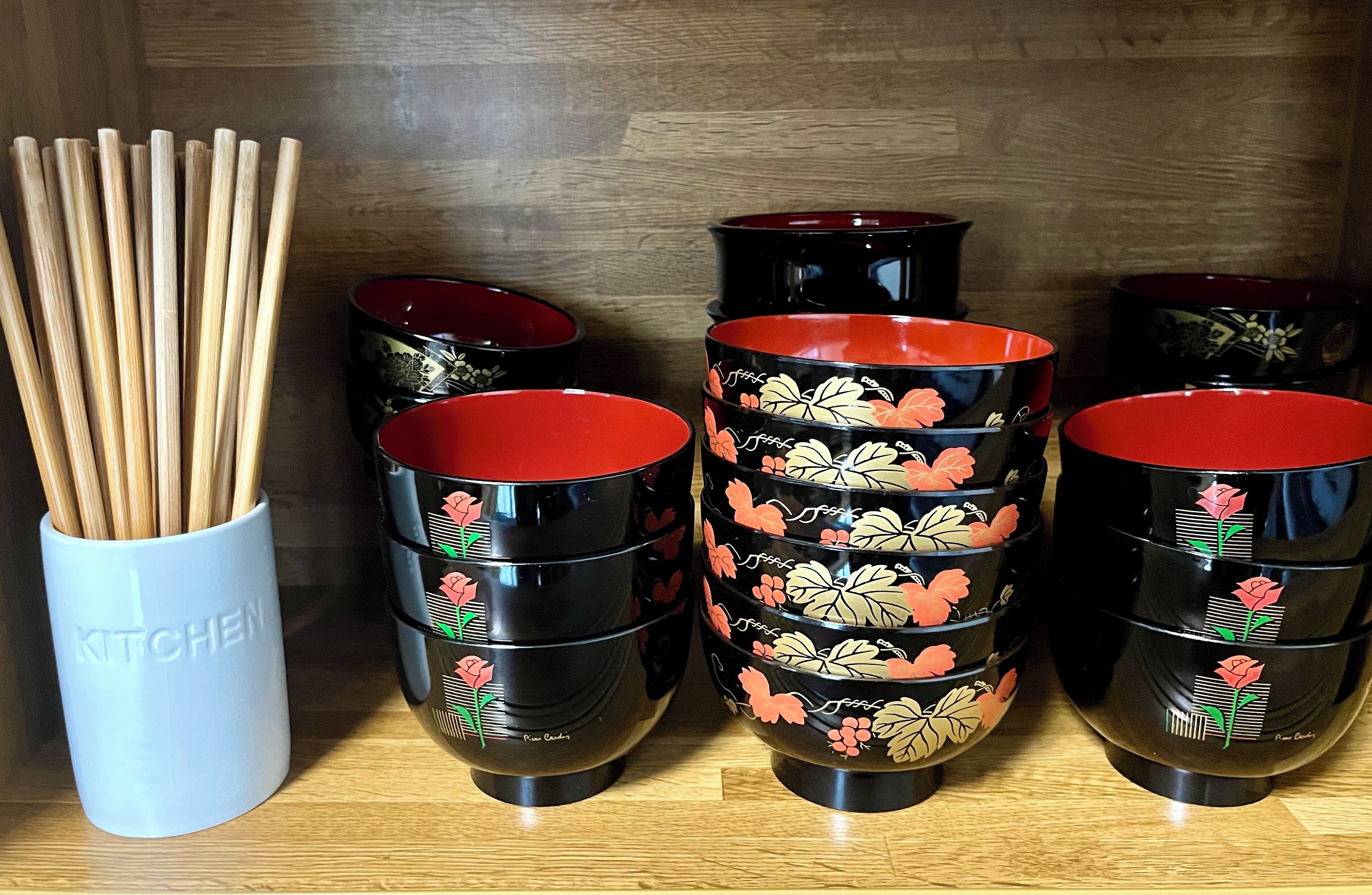 Conjunto de tigelas pretas e vermelhas com desenhos florais, um recipiente branco com a etiqueta "KITCHEN" e um recipiente de madeira com hashis.