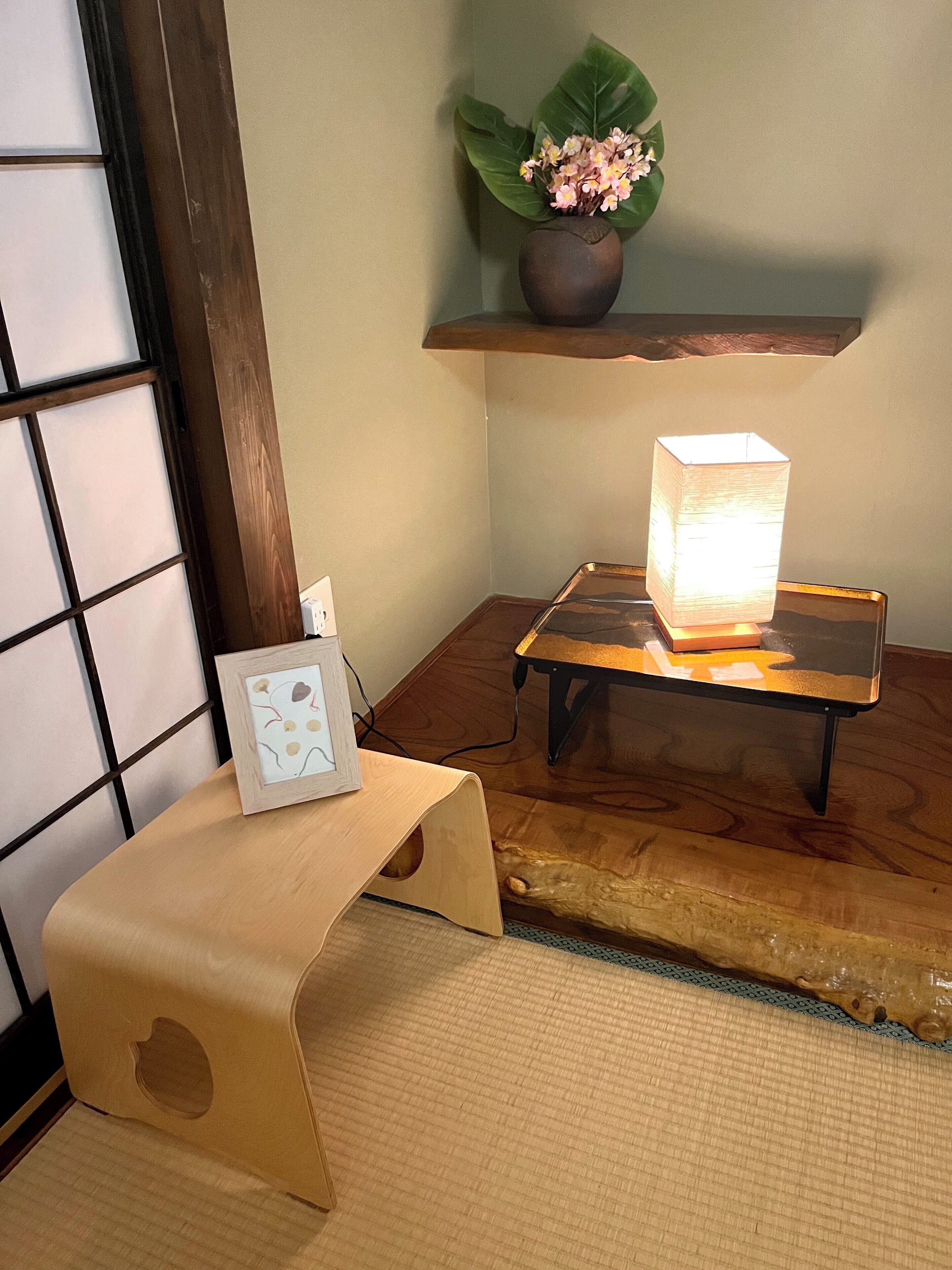 Une pièce de style japonais traditionnel, avec un sol recouvert de tatamis, une basse table en bois sur laquelle se trouve une lampe, une petite étagère avec une plante et un tableau encadré posé sur un petit tabouret.
