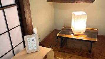 Une pièce de style japonais traditionnel, avec un sol recouvert de tatamis, une basse table en bois sur laquelle se trouve une lampe, une petite étagère avec une plante et un tableau encadré posé sur un petit tabouret.