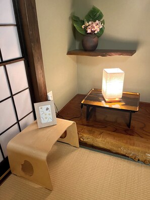 A traditional Japanese room with tatami flooring, a low wooden table with a lamp, a small shelf with a plant, and a framed picture on a low stool. - Mitsuba House (Annaka)