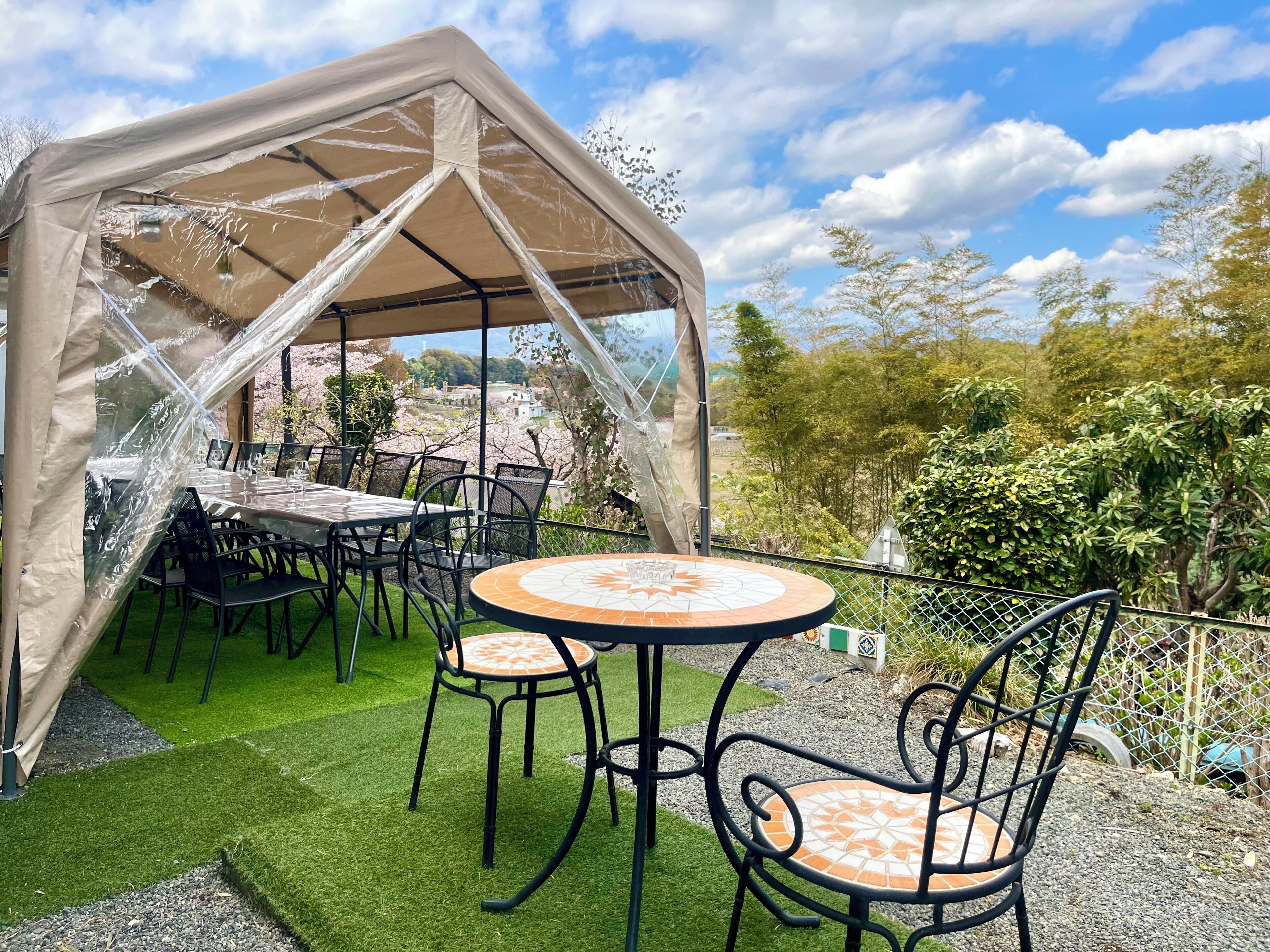 Een overdekt terras met tafels en stoelen, omgeven door groen en een heldere lucht.