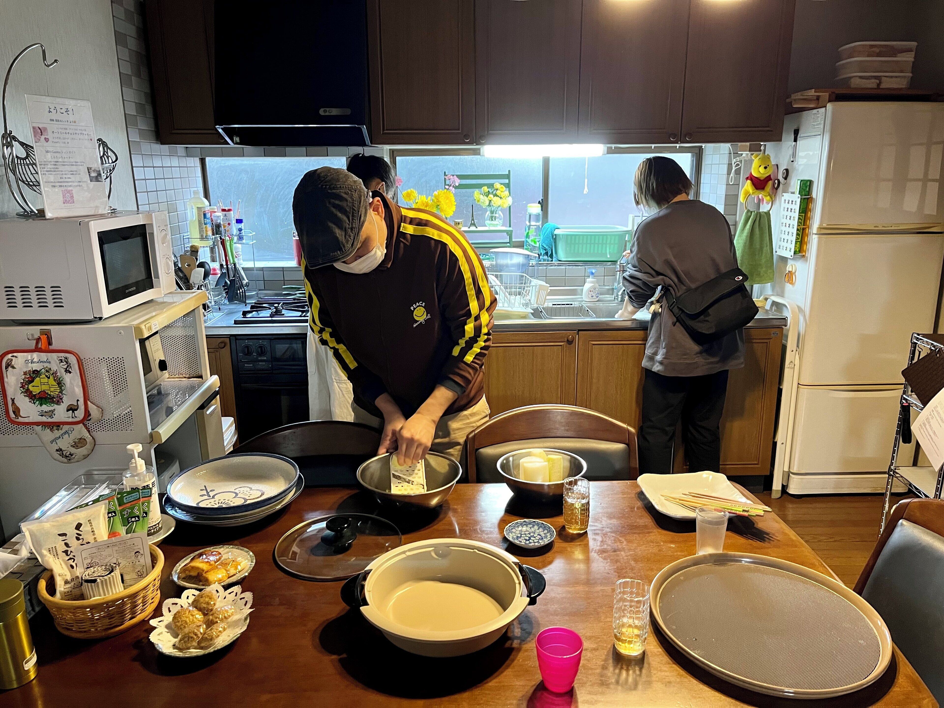 Una cocina con armarios de madera, un microondas y un refrigerador. Hay varios utensilios de cocina en la encimera, incluyendo cuencos, una cesta y una botella.