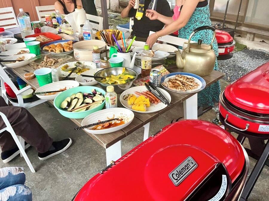 Mesa de jantar comunitária com diversos pratos e um churrasqueira Coleman.