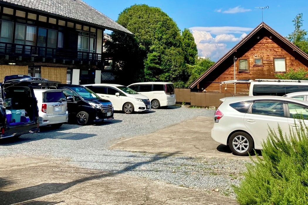 Un estacionamiento con varios autos, un edificio de techo inclinado y una estructura de madera con techo triangular.