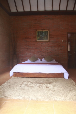 Een kamer met een bakstenen muur, een bed met witte beddengoed en twee kussens, een schilderij aan de muur en een deur aan de rechterkant.