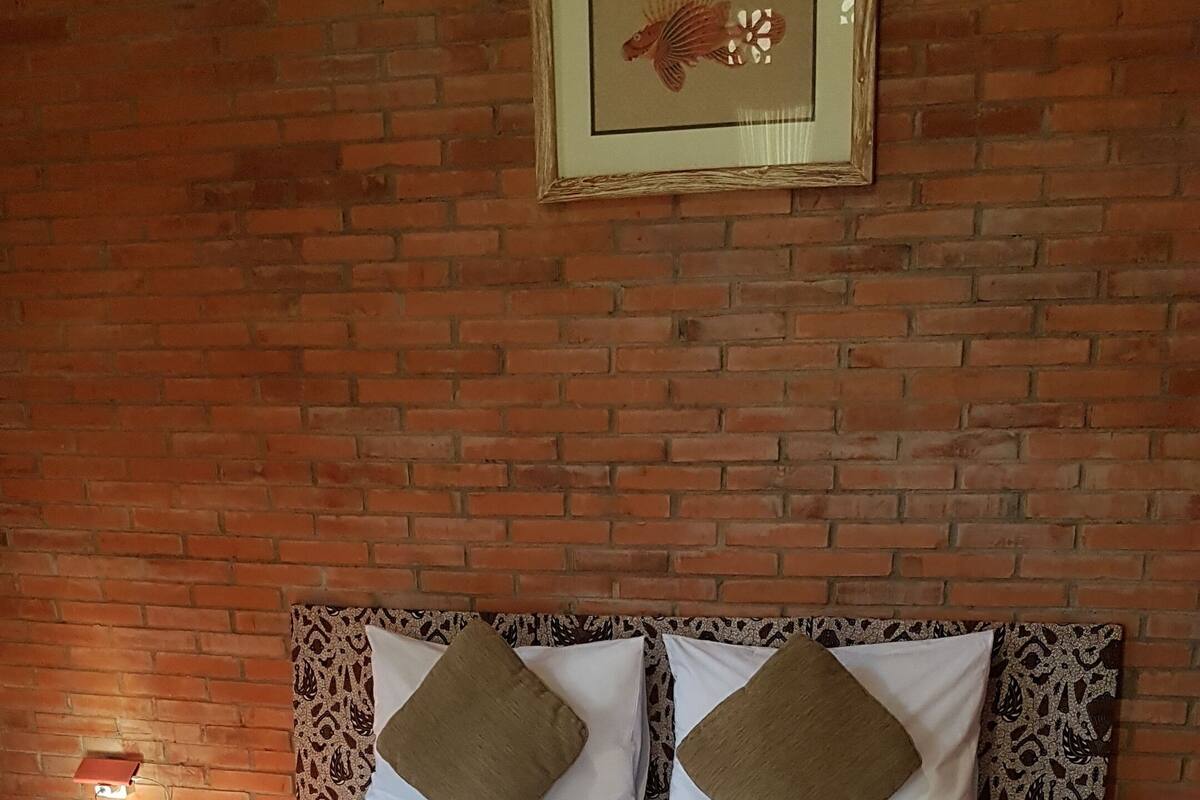 A bed with a patterned bedspread, two pillows, and a framed picture on a brick wall.