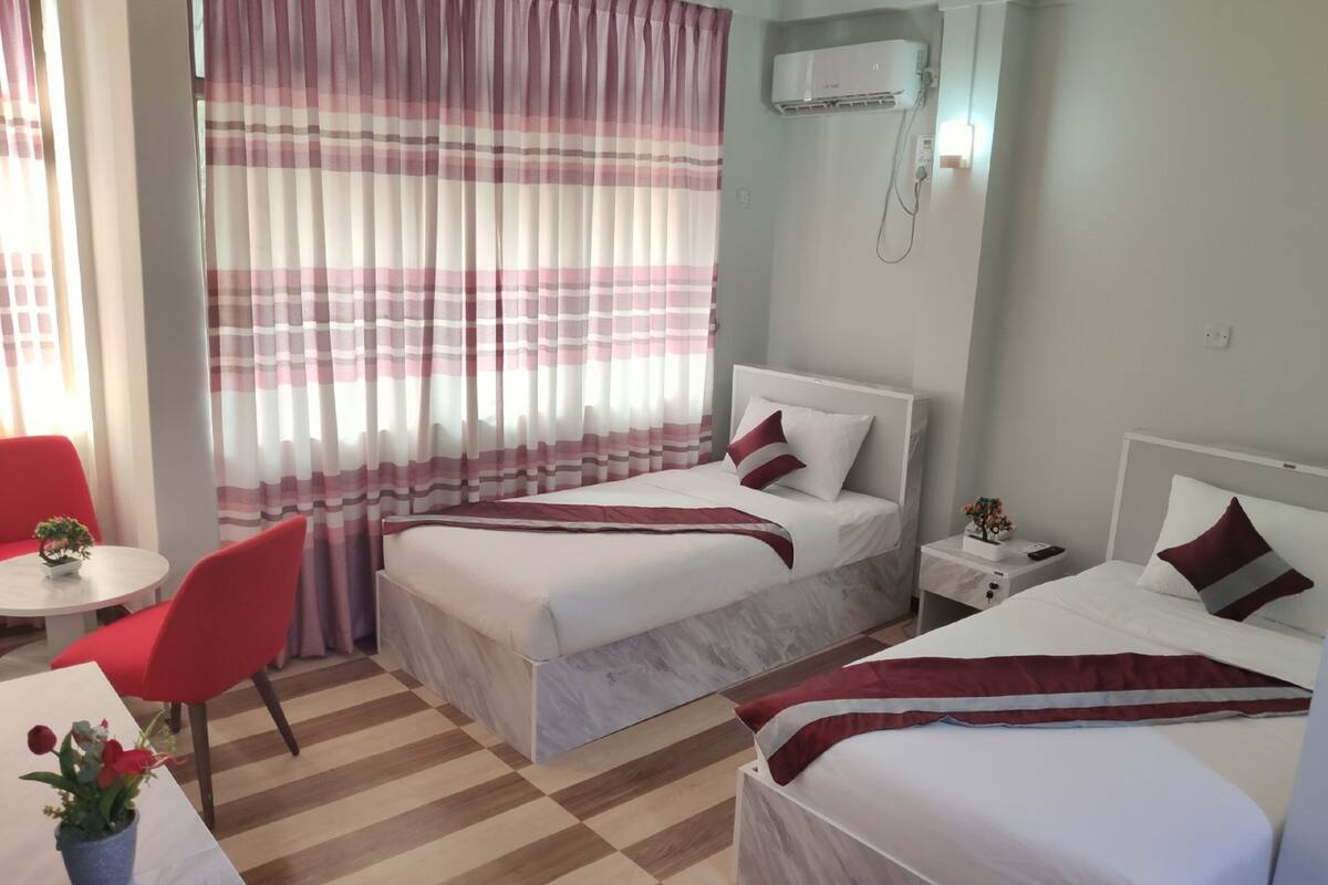 A hotel room with two beds, a red chair, a small table with a plant, and a window with striped curtains.