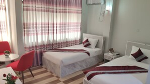 Ein Hotelzimmer mit zwei Betten, einem roten Sessel, einem kleinen Tisch mit Pflanze und einem Fenster mit gestreifter Vorhang.