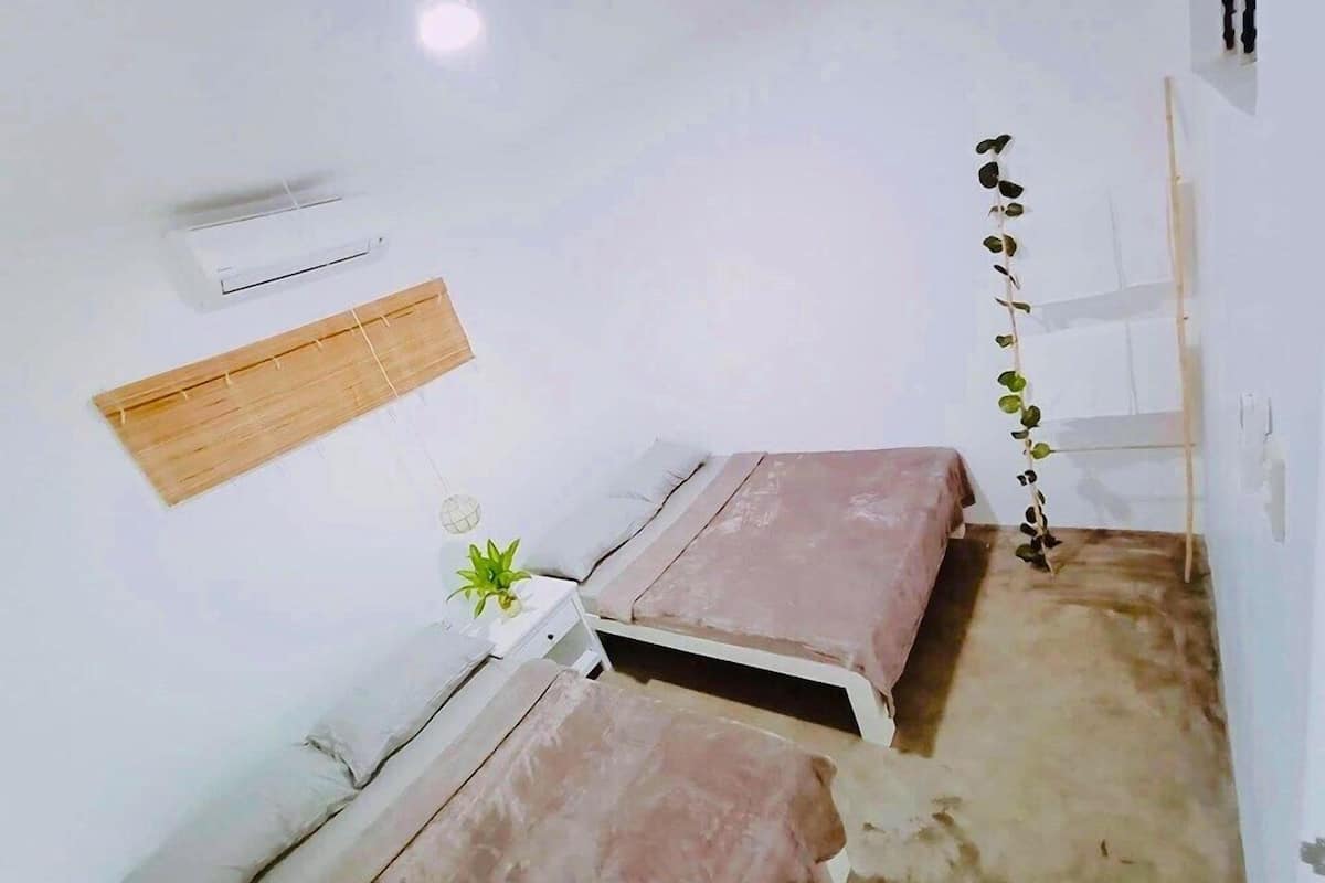 Una habitación pequeña y funcional con dos camas, aire acondicionado de pared, una pequeña planta en maceta y un estante de madera.