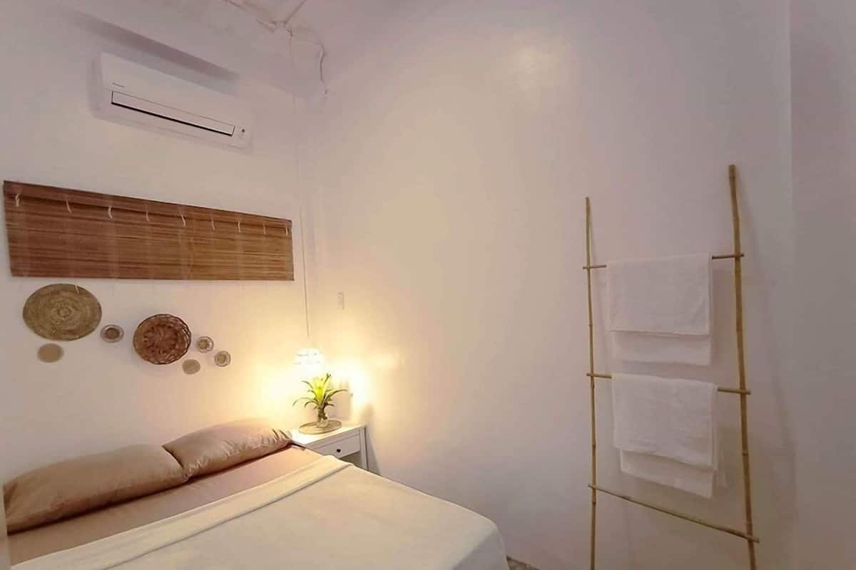 Un dormitorio con una cama, una mesita de noche con lámpara, aire acondicionado de pared y un perchero de bambú.