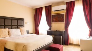 Ein Hotelzimmer mit einem großen Bett, einem Schreibtisch und einem Sessel. Ein Fenster mit roten Vorhängen und eine Klimaanlage an der Wand sind zu sehen.