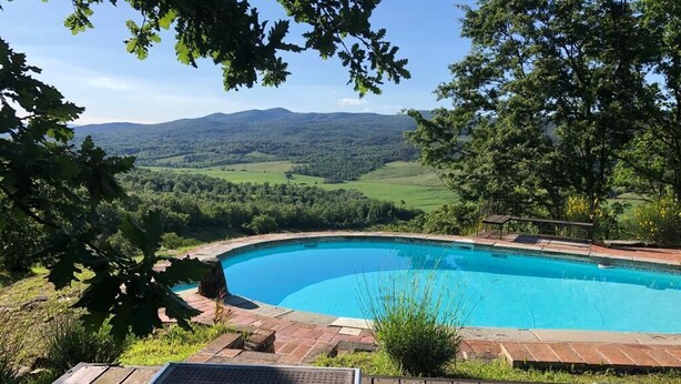 Una piscina con cubierta de madera y sillas de descanso, rodeada de vegetación y vistas a colinas distantes.