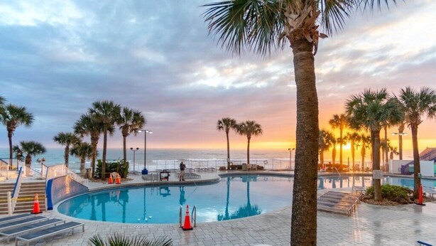 A pool area with palm trees, a curved pool, and a view of the sunset over the ocean. - Daytona Beach | Beach & Pool Access | 2BR Deluxe (Daytona)